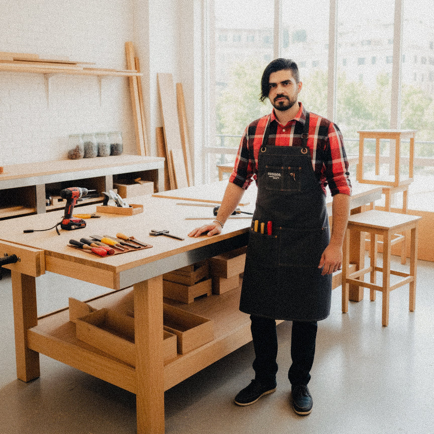 Man in a workshop wearing a black apron and standing next to a wooden workbench with tools.