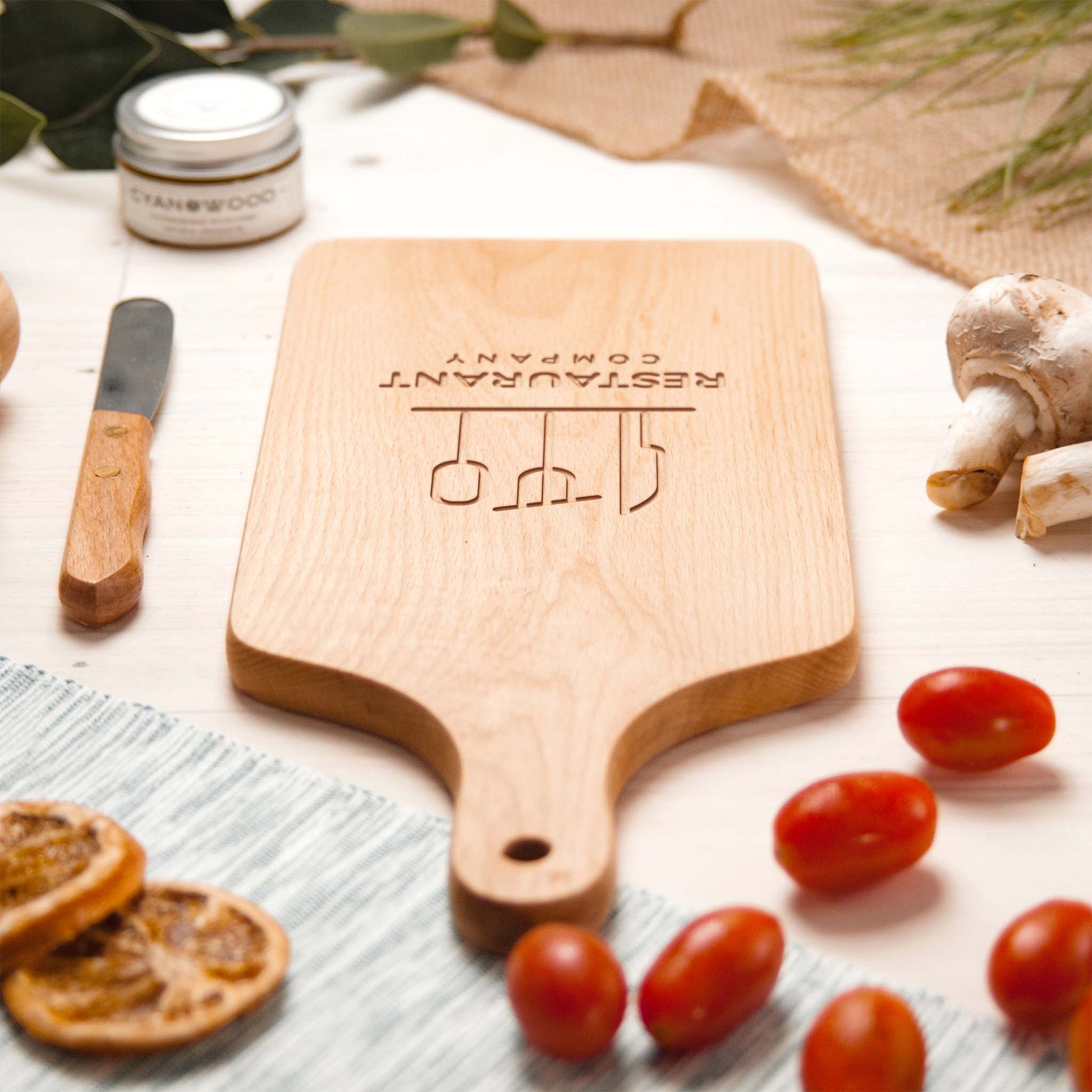 Wooden cutting board with 'Restaurant Company' engraving on a table with tomatoes and herbs.