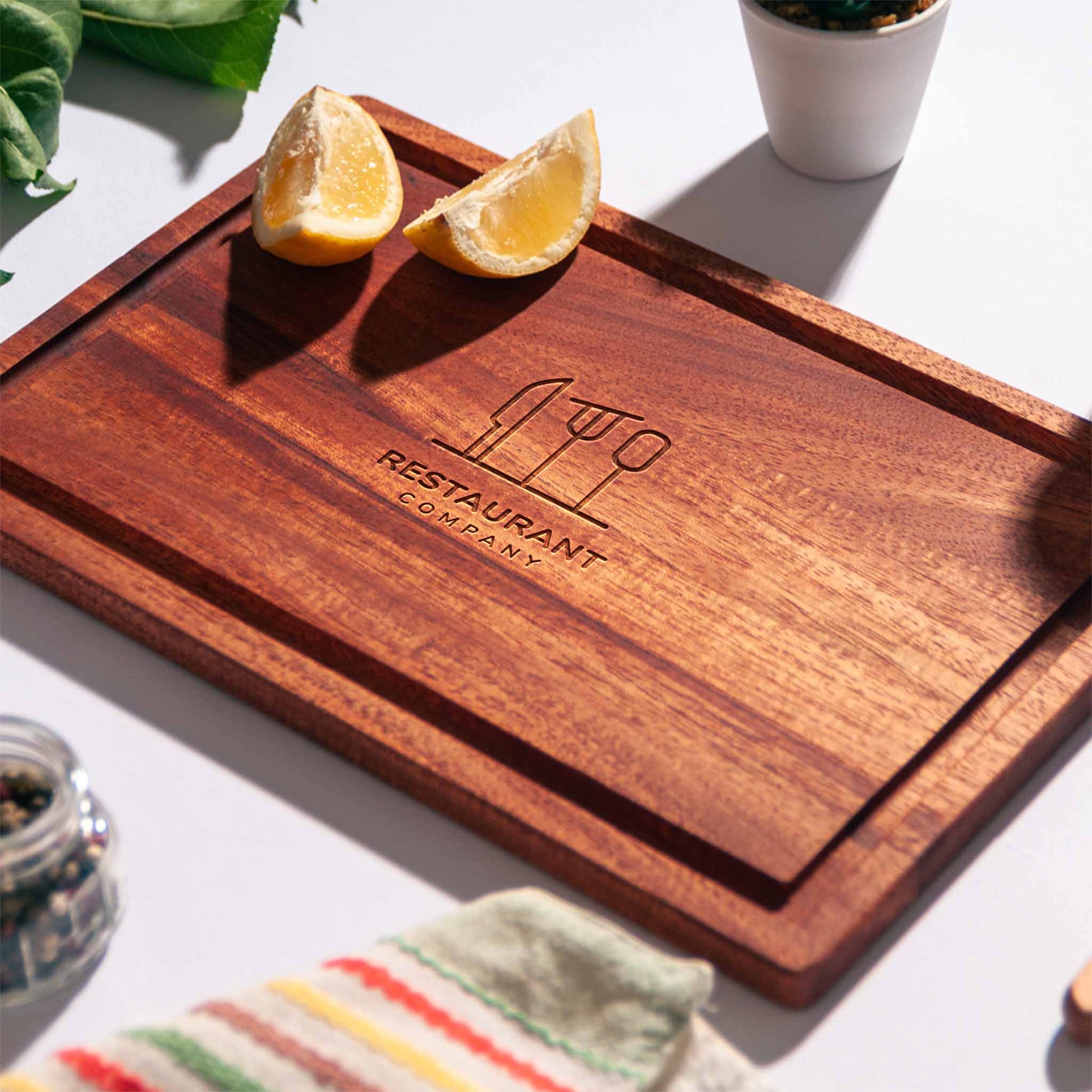 Rectangular mahogany cutting board with juice groove and engraved restaurant logo, styled with lemon halves, pepper mill and napkin.