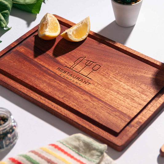 Rectangular mahogany cutting board with juice groove and engraved restaurant logo, styled with lemon halves, pepper mill and napkin.