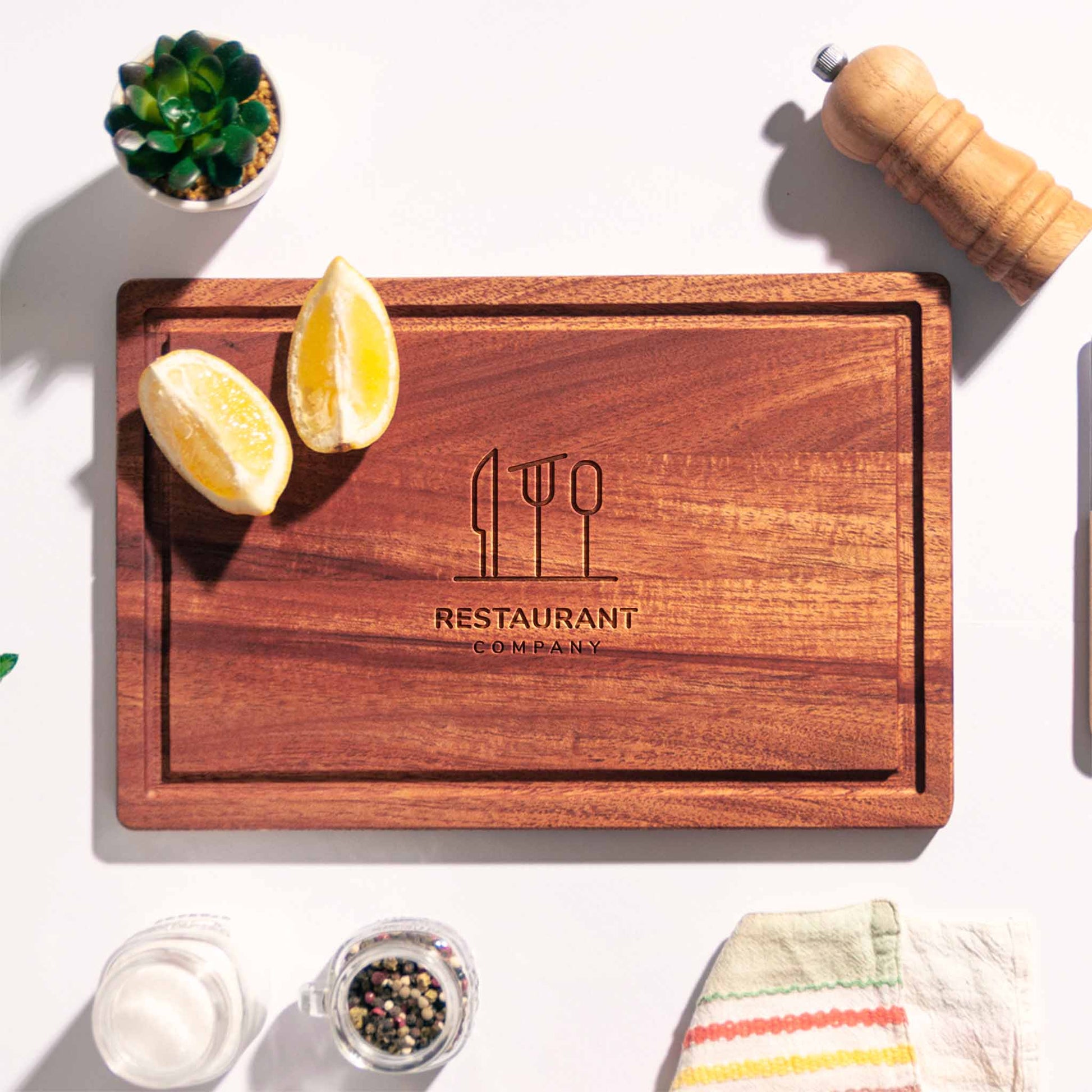 Rectangular mahogany cutting board with juice groove and engraved restaurant logo, styled with lemon halves, pepper mill and napkin.