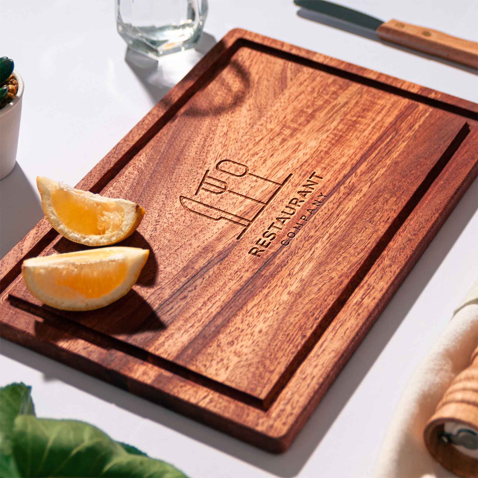 Rectangular mahogany cutting board with juice groove and engraved restaurant logo, styled with lemon halves, pepper mill and napkin.